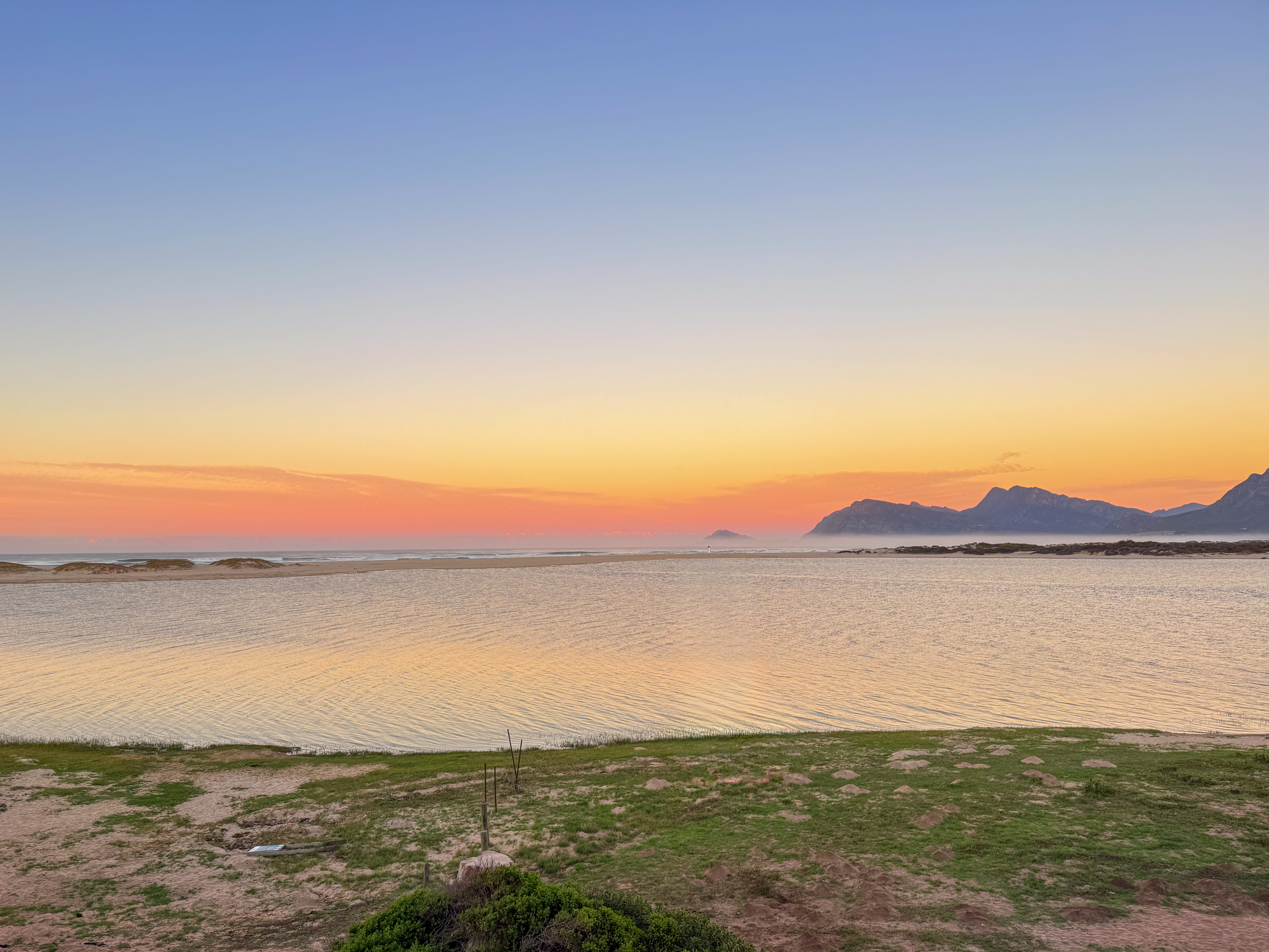 Pink and orange sunrise over the Kleinrivier Lagoon at Fisherhaven, with the lagoon mouth and mountains of Walker Bay visible in the distance